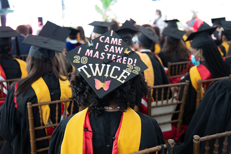 A student seated with a cap decorated to read "I CAME I SAW I MASTERED TWICE, 20 / 22"